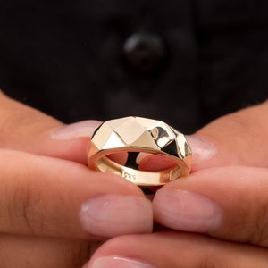 Könnte beinhalten: Ein goldener Ring mit geometrischem Design. Der Ring ist aus glattem, glänzendem Metall und hat ein breites Band.