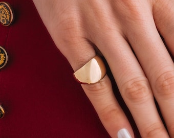 May include: A gold ring with a rounded, slightly curved top. The ring is worn on the middle finger of a hand with white nail polish.