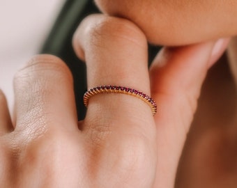 Könnte beinhalten: Ein goldener Ring mit einem Band aus kleinen, dunkelroten Edelsteinen. Der Ring befindet sich an einem Finger, und die Person stützt ihr Kinn auf ihre Hand. Der Ring ist ein zarter Schmuck.