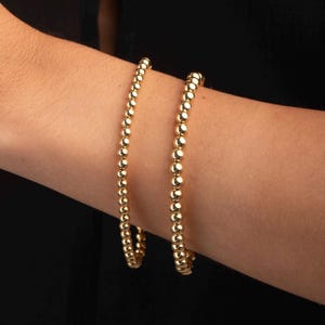 Puede incluir: Dos pulseras doradas con cuentas se muestran en un brazo sobre un fondo negro. Las pulseras están hechas de pequeñas cuentas redondas doradas ensartadas. Las pulseras se usan juntas en el brazo.