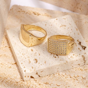 May include: Two gold rings on a white textured surface. One ring is a simple band with a flat top, the other ring has a square top with small clear stones.