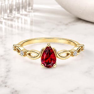 Könnte beinhalten: Ein Goldring mit einem birnenförmigen roten Edelstein in der Mitte, flankiert von zwei kleinen klaren Steinen auf jeder Seite. Das Band hat ein kompliziertes, durchbrochenes Design und ist mit der Zahl 585 auf der Innenseite geprägt.