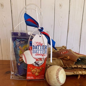 May include: A clear tote bag filled with snacks and a baseball-themed gift tag that reads "Hey Batter Batter Swing". A baseball, a baseball glove, and a bag of caramel popcorn are also included.