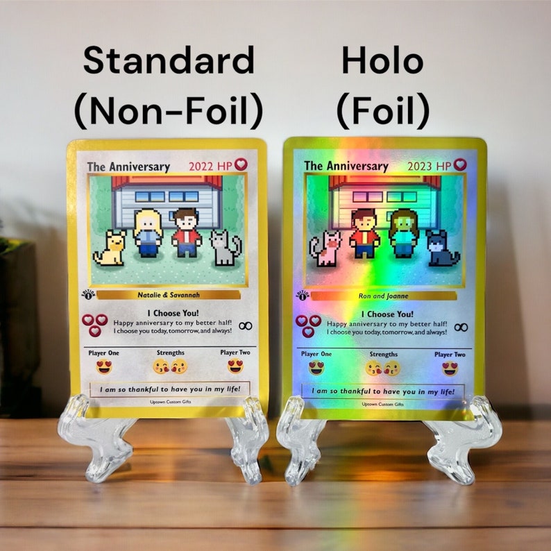 May include: Two versions of a personalized card game featuring a pixelated image of a couple and two cats in front of a house. The card on the left is a standard card with a yellow border. The card on the right is a holographic card with a rainbow border. Both cards have the text "The Anniversary" and a year, 2022 or 2023, and the text "I Choose You!" with a heart symbol.