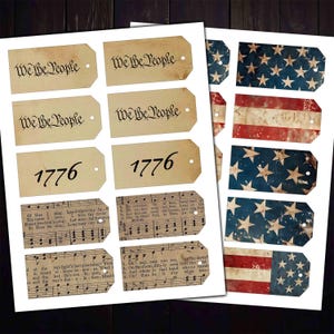 May include: A collection of vintage-style paper tags with patriotic themes. Some tags feature the text "We the People" and "1776" in a classic font. Other tags display the American flag or sheet music. The tags are aged with a distressed look.