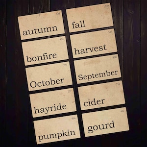 May include: Ten vintage-style cards with fall-themed words: autumn, fall, harvest, bonfire, October, hayride, September, cider, pumpkin, and gourd. The cards are beige with black text, arranged on a dark wood surface.