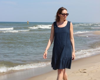 May include: A sleeveless navy blue dress with a ruffled hem is worn on a sandy beach. The dress has a scoop neckline. The person is wearing sunglasses and walking near the ocean under a blue sky.