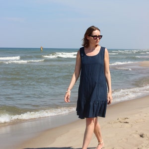 May include: A sleeveless navy blue dress with a ruffled hem is worn on a sandy beach. The dress has a scoop neckline. The person is wearing sunglasses and walking near the ocean under a blue sky.