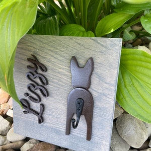 May include: A wooden sign with a gray wash finish featuring a black silhouette of a dog's rear end with a hook attached. The dog's name "Libby" is written in black cursive lettering on the sign.