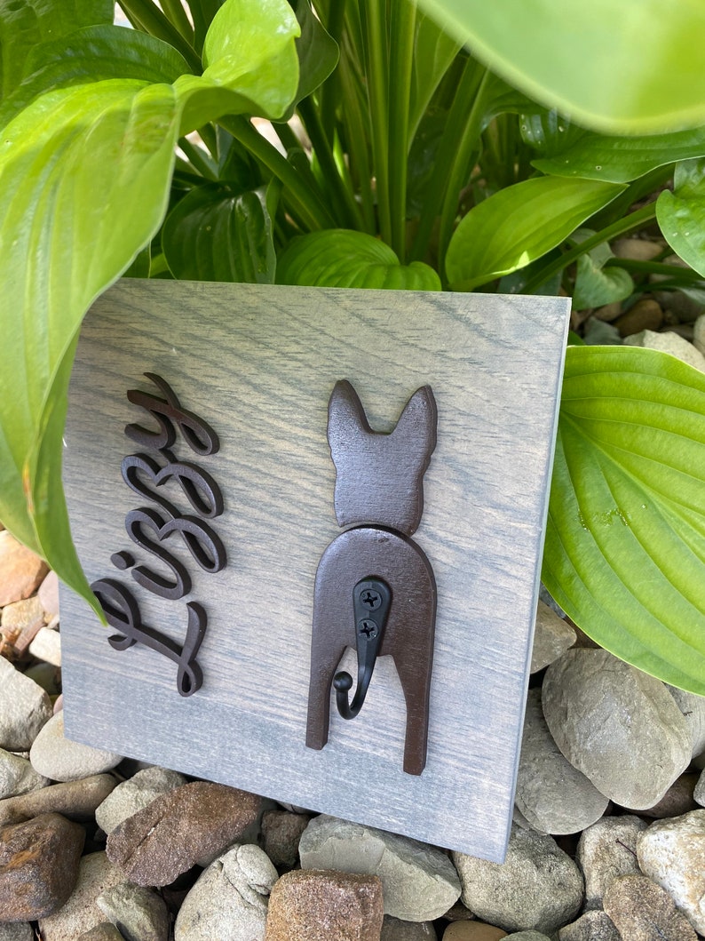May include: A wooden sign with a gray wash finish featuring a black silhouette of a dog's rear end with a hook attached. The dog's name "Libby" is written in black cursive lettering on the sign.