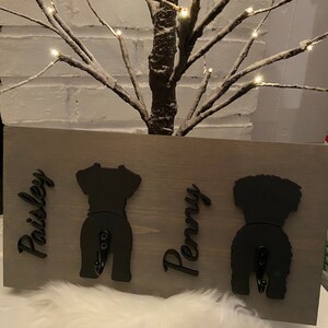May include: A gray wooden sign with two black dog silhouettes, each with a hook. The names "Paisley" and "Penny" are written on the sign.