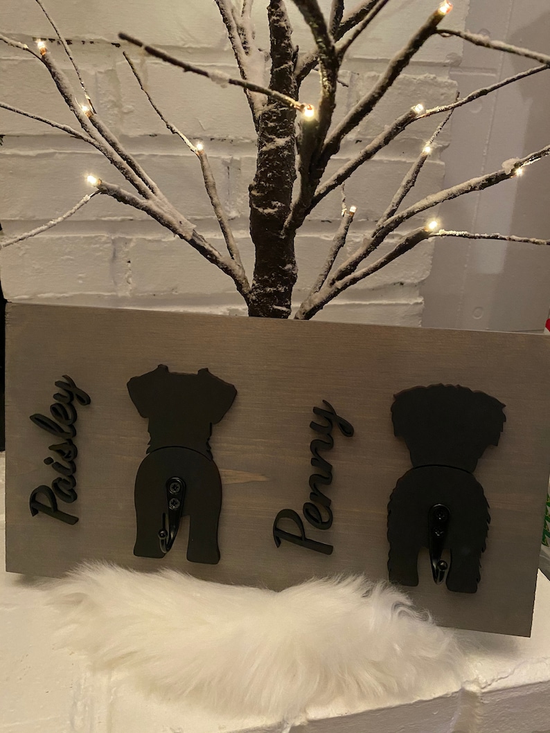 May include: A gray wooden sign with two black dog silhouettes, each with a hook. The names "Paisley" and "Penny" are written on the sign.