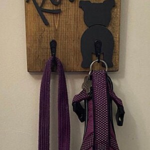 May include: A wooden dog leash holder with the name "Rey" written in black cursive lettering. A black silhouette of a dog's rear end is on the right side of the holder. A purple leash and a purple dog harness are hanging from the hooks.