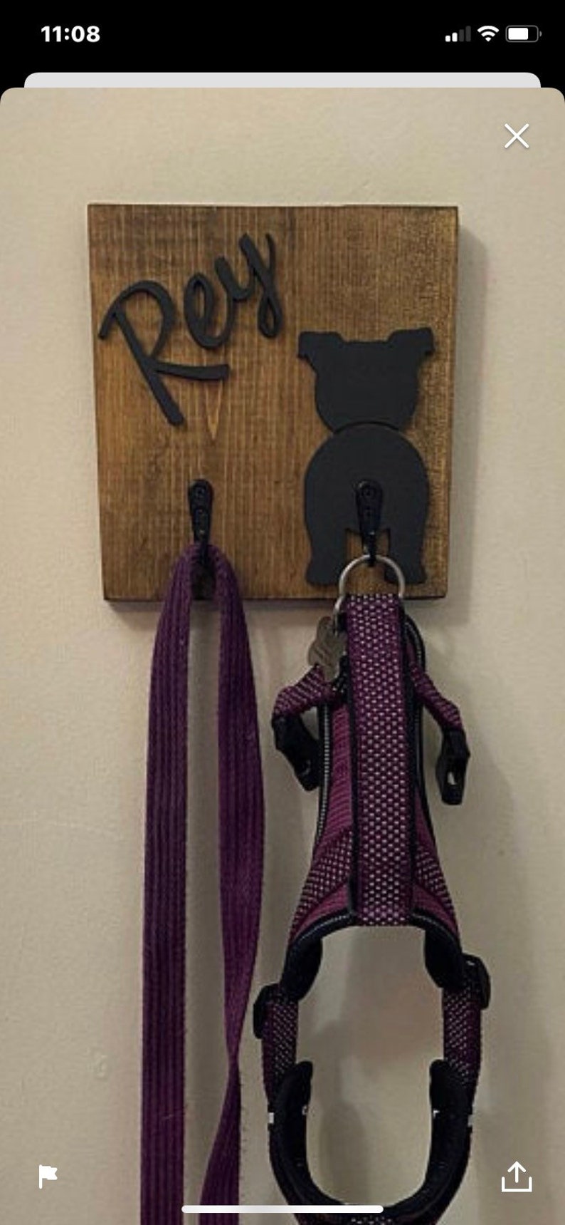 May include: A wooden dog leash holder with the name "Rey" written in black cursive lettering. A black silhouette of a dog's rear end is on the right side of the holder. A purple leash and a purple dog harness are hanging from the hooks.