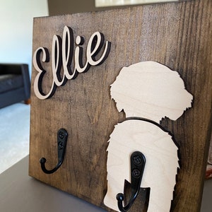 May include: A wooden dog leash holder with the name "Ellie" in black script. The holder features a cutout of a dog's rear end and two black metal hooks.