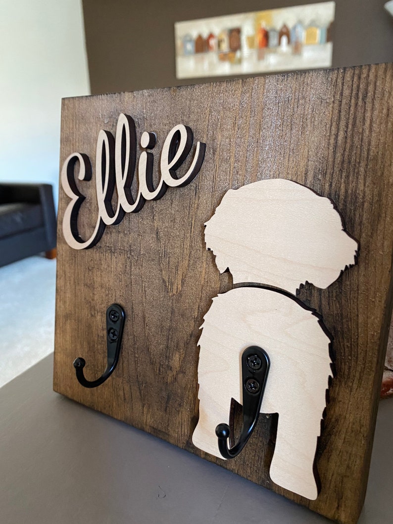 May include: A wooden dog leash holder with the name "Ellie" in black script. The holder features a cutout of a dog's rear end and two black metal hooks.