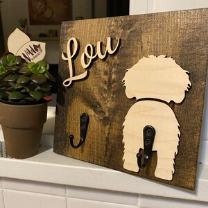 May include: A wooden sign with the name "Lou" written in cursive on it. The sign has two black metal hooks, one shaped like a dog's tail. The sign is dark brown with a light brown dog silhouette.