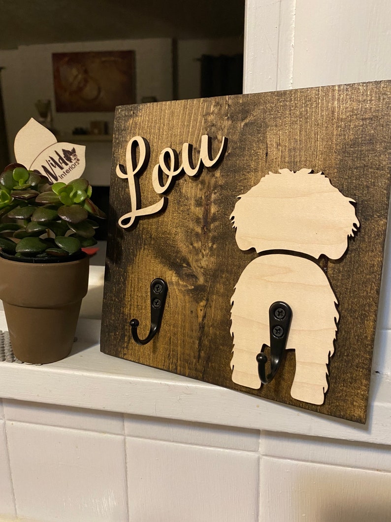 May include: A wooden sign with the name "Lou" written in cursive on it. The sign has two black metal hooks, one shaped like a dog's tail. The sign is dark brown with a light brown dog silhouette.