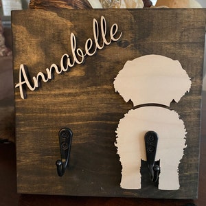 May include: A wooden dog leash holder with two hooks. The holder is painted brown and has a white silhouette of a dog's rear end. The name "Annabelle" is written in cursive on the holder.