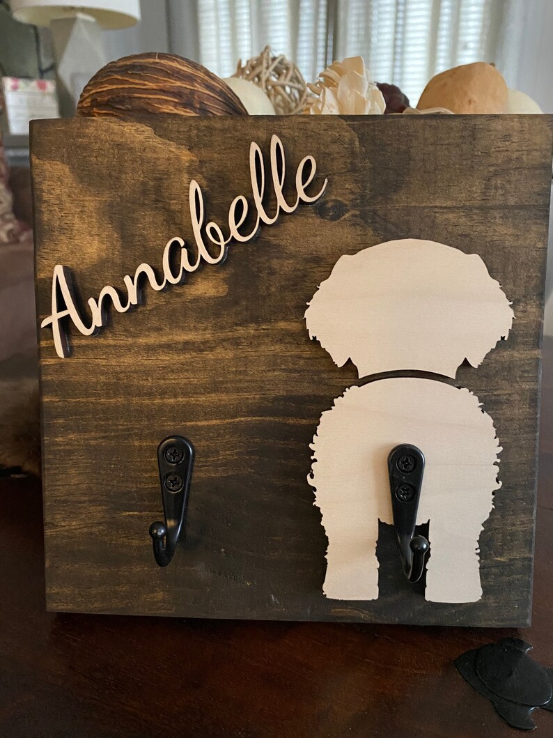 May include: A wooden dog leash holder with two hooks. The holder is painted brown and has a white silhouette of a dog's rear end. The name "Annabelle" is written in cursive on the holder.