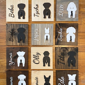 May include: A wall mounted dog leash holder with 15 wooden plaques. Each plaque features a silhouette of a dog's rear end with a hook attached. The plaques are painted in various shades of brown and white. The names of the dogs are written on each plaque, including Luna, Yuna, Brody, Beba, Trickle, Louie, Olly, Piper, Tycho, Charlie, Ducy, Like, Tito, Sophie, and Lizzy.