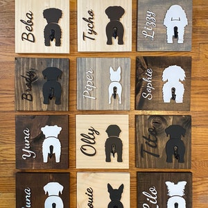 May include: A wall mounted dog leash holder with 15 wooden plaques. Each plaque features a silhouette of a dog's rear end with a hook attached. The plaques are painted in various shades of brown and white. The names of the dogs are written on each plaque, including Luna, Yuna, Brody, Beba, Trickle, Louie, Olly, Piper, Tycho, Charlie, Ducy, Like, Tito, Sophie, and Lizzy.