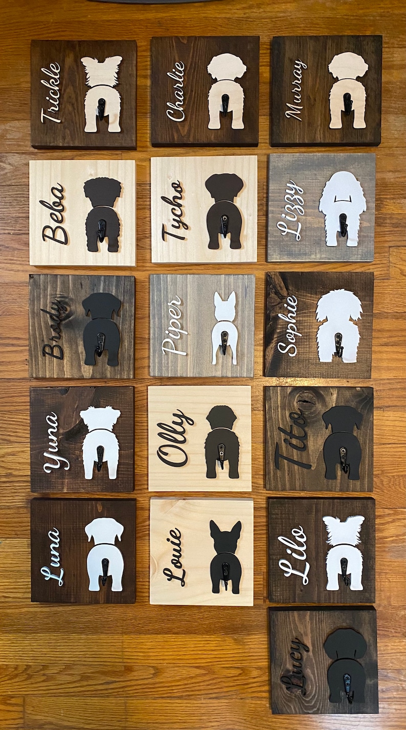 May include: A wall mounted dog leash holder with 15 wooden plaques. Each plaque features a silhouette of a dog's rear end with a hook attached. The plaques are painted in various shades of brown and white. The names of the dogs are written on each plaque, including Luna, Yuna, Brody, Beba, Trickle, Louie, Olly, Piper, Tycho, Charlie, Ducy, Like, Tito, Sophie, and Lizzy.