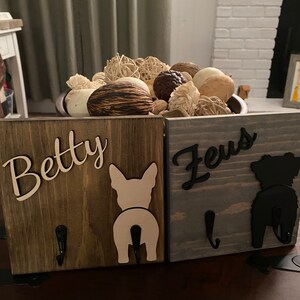 May include: Two wooden dog leash hooks with the names "Betty" and "Zeus" written on them. Each hook has a silhouette of a dog's rear end, one white and one black.