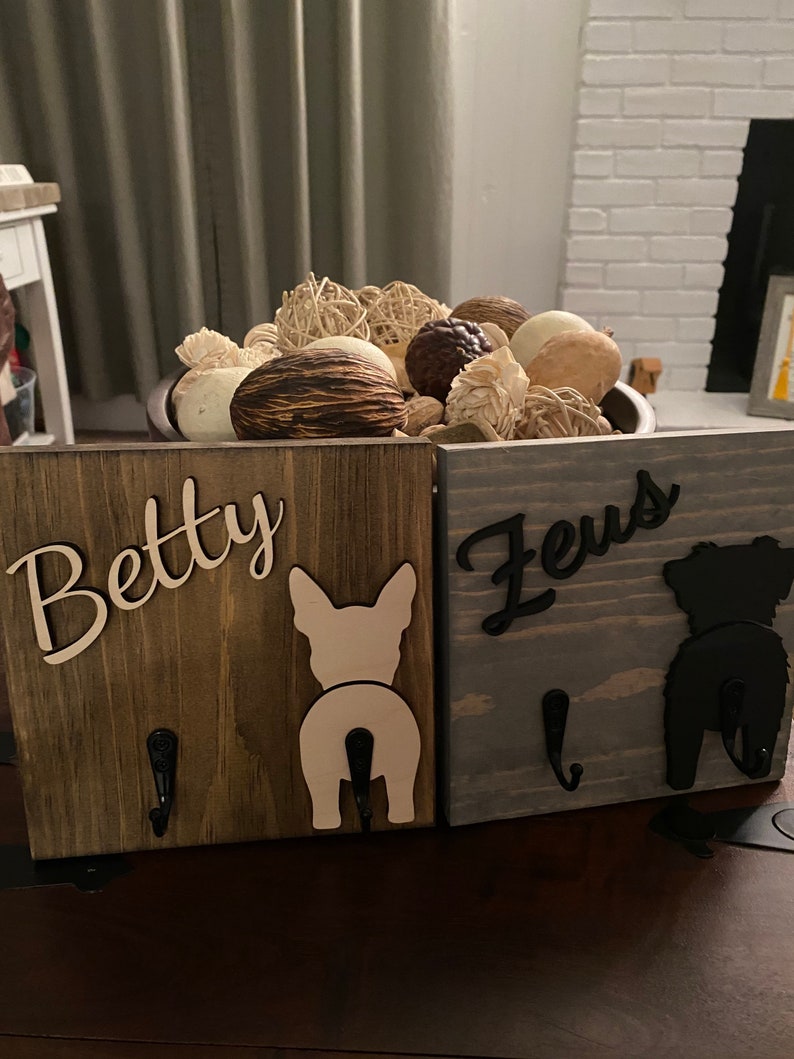 May include: Two wooden dog leash hooks with the names "Betty" and "Zeus" written on them. Each hook has a silhouette of a dog's rear end, one white and one black.