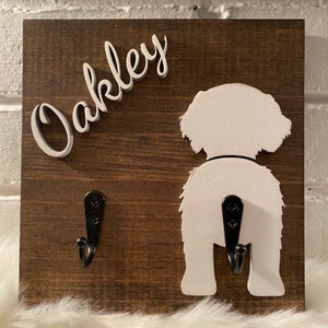 May include: A wooden dog leash holder with two hooks. The holder is painted brown and features a white silhouette of a dog's rear end with the name "Oakley" written in white on the top left corner.