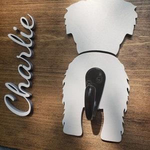 May include: Wooden sign with a white silhouette of a dog's rear end with a black hook attached. The name "Charlie" is written in white on the left side of the sign.