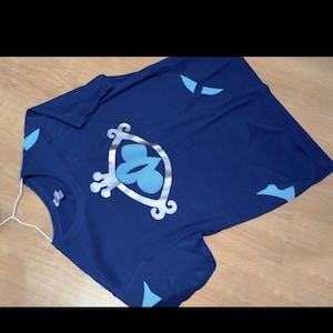 Peut inclure: Un t-shirt bleu marine avec un motif argenté et bleu clair. Le motif représente un œil stylisé avec un papillon à l'intérieur.