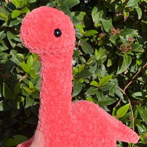 May include: A coral-coloured, plush dinosaur toy with a long neck, small legs, and a tail. The dinosaur has a round head with a single black eye. The toy is made of a soft, textured material, and is set against a background of green leaves.