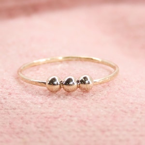 May include: A delicate gold ring featuring three small, shiny spheres centered on the band. The ring has a hammered texture and a thin band. The ring is displayed on a soft pink surface.