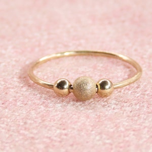 Può includere: Un delicato anello d'oro con tre sfere decorative. La sfera centrale ha una finitura strutturata e opaca, mentre le due sfere laterali sono lisce. L'anello è posto su uno sfondo rosa tenue.