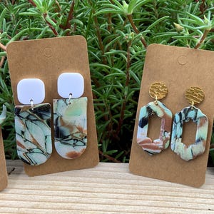 May include: Four pairs of handmade polymer clay earrings. The earrings feature different shapes and designs, including a skull, a rectangle, a square, and a freeform shape. The earrings are all painted in a turquoise and brown marbled pattern.
