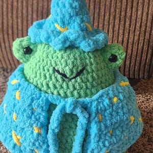 May include: A handmade green crochet frog wearing a blue wizard hat and cape, adorned with yellow star and crescent moon accents. The frog has black eyes and a smiling mouth. A whimsical, soft toy.
