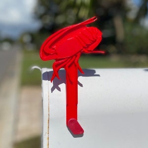 May include: A bright red lobster mailbox flag. The flag is made of a solid material and is attached to a white mailbox. The lobster design is detailed and stands out against the mailbox.