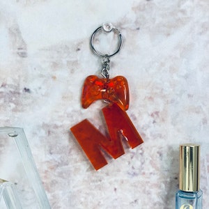May include: Orange resin keychain featuring a letter M and a video game controller charm. The keychain is attached to a silver key ring.