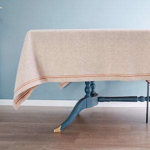 May include: A beige linen tablecloth with a thin, double rust-colored stripe along the edge covers a blue table with gold-tipped legs. A chair with a floral print seat and back is partially visible.