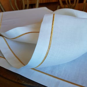 Può includere: Tovaglia di lino bianca con bordatura ricamata in oro. Il tessuto è piegato, mettendo in risalto la trama e le cuciture dettagliate. Questa tovaglia è probabilmente per un tavolo da pranzo.