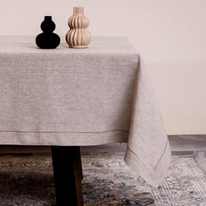 May include: A beige linen tablecloth with a gold trim, draped over a dark wooden table. Two decorative vases sit on the table: a black, rounded vase and a beige, ribbed vase. The tablecloth has a simple, elegant design.