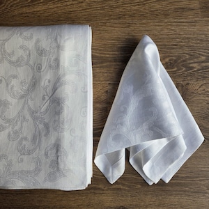 May include: A white damask tablecloth and matching napkin set. The tablecloth is folded, displaying a detailed, light grey floral pattern. The napkin is folded in a triangle, showing the same design. The set is on a wooden surface.