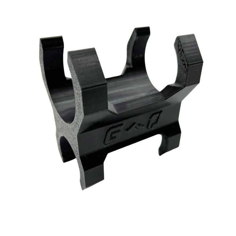 Może przedstawiać: Czarny wspornik drukowany w 3D z zakrzywioną g&oacute;rą i czterema nogami. Na wsporniku wygrawerowano litery "G < P". Wspornik jest przeznaczony do trzymania przedmiotu.