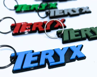 May include: Keychains with the word "TERYX" in various colors, including blue, green, red, and orange. Each keychain has a black border and a silver keyring attached. The keychains are arranged on a white surface.