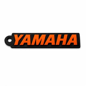Pu&ograve; includere: Un portachiavi rettangolare nero con la scritta "YAMAHA" in lettere arancioni brillanti. Il portachiavi ha un piccolo foro circolare sul lato sinistro per l'attacco. Lo sfondo &egrave; bianco.