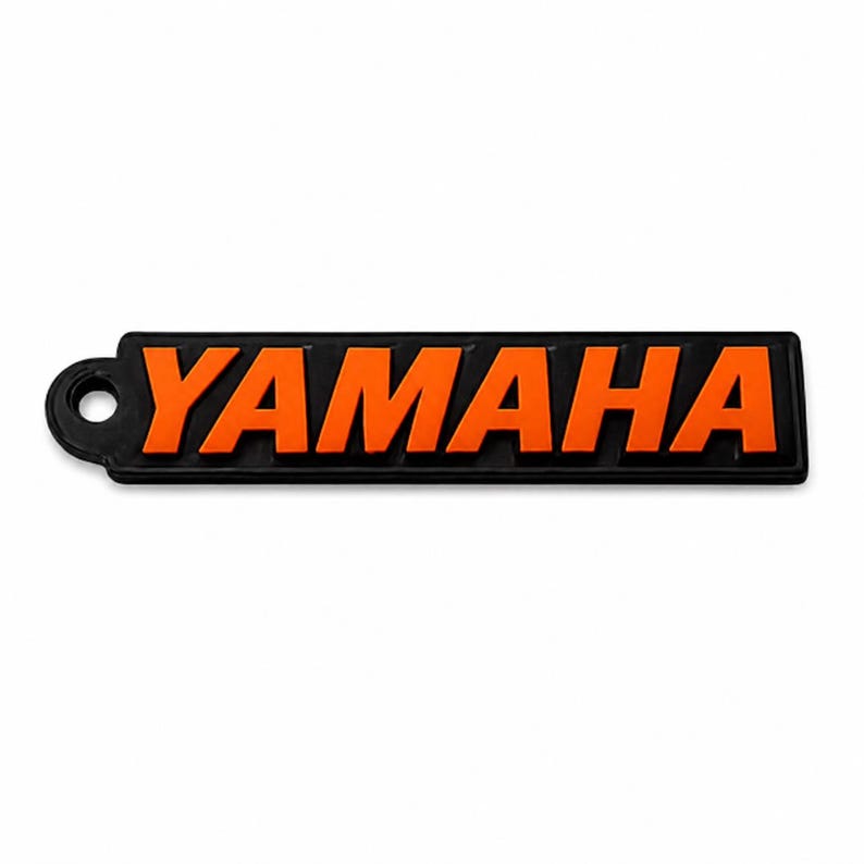 Pu&ograve; includere: Un portachiavi rettangolare nero con la scritta "YAMAHA" in lettere arancioni brillanti. Il portachiavi ha un piccolo foro circolare sul lato sinistro per l'attacco. Lo sfondo &egrave; bianco.