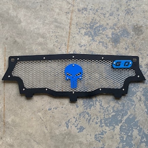 May include: Black metal grille with a blue skull and G-O logo. The grille is made of a mesh material and has a black powder coat finish.