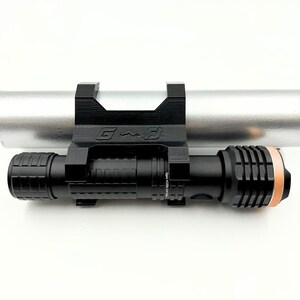 Może przedstawiać: Czarna latarka zamontowana na czarnym wsporniku, przymocowana do srebrnego cylindrycznego przedmiotu. Latarka ma teksturowany uchwyt i miedziany pierścień na końcu. Na wsporniku widnieją litery "G" i "D".