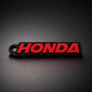 May include: A black and red Honda keychain. The word "HONDA" is in red, bold letters on a black rectangular background. The keychain has a small hole on the left side for attachment.